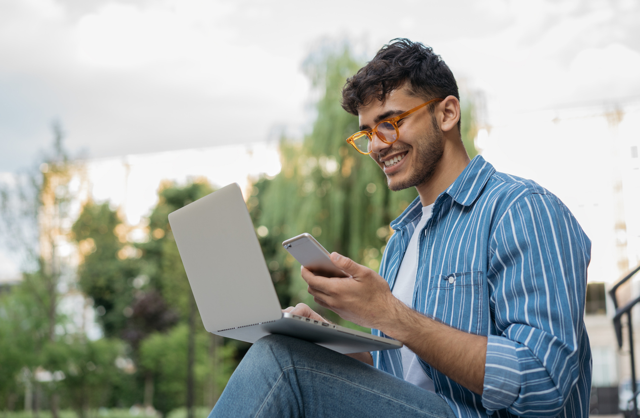 Young handsome Indian man using laptop computer, mobile phone, working freelance project online, sitting outdoors.jpg