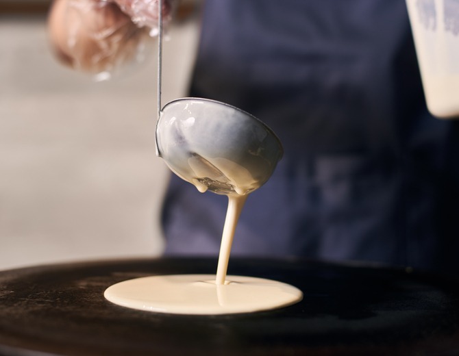 Woman chef pour dough by hands with ladle on cooktop. Cheese, tomato slice in plate on blurred background. Front view.