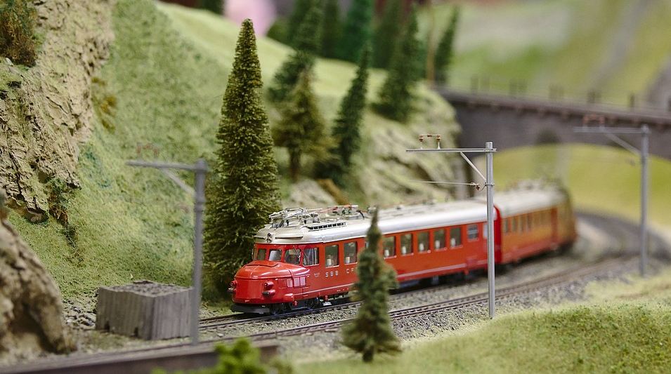 a red model train passing through a model railroad with terrains and pine trees in the background