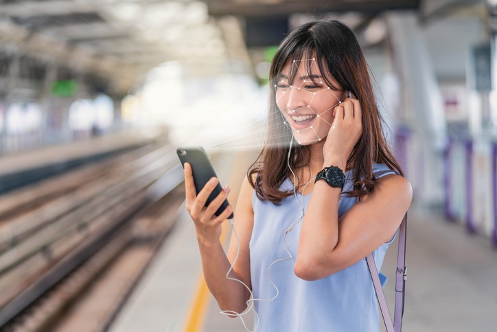 Asian woman using face recognition via smart mobile phone and listening the music at Railroad Station Platform, Biometric Verification and artificial intelligence concept