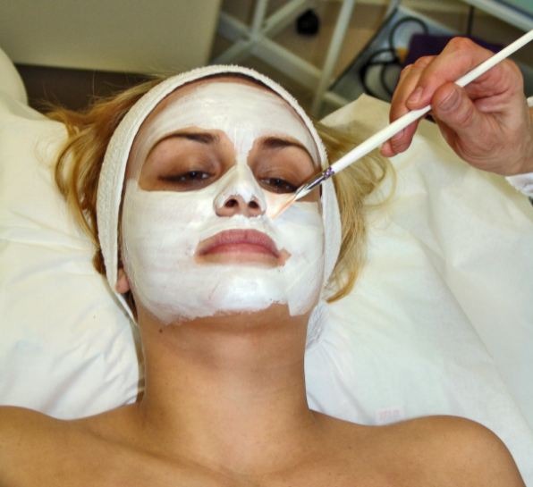 girl laying down, white face mask applied on the skin, hand holding a white face brush
